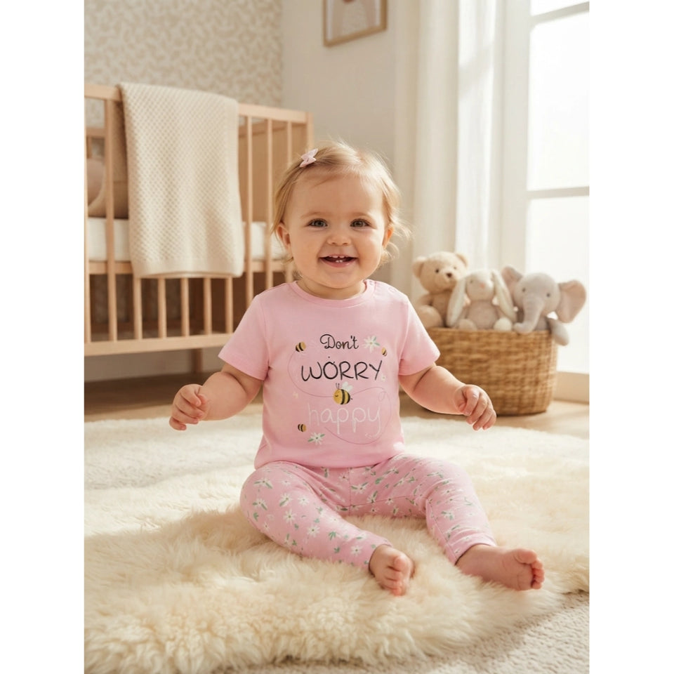 A baby girl sitting on a fluffy cream rug in a bright nursery, wearing a light pink short-sleeve pajama top with 'Don't WORRY happy' printed on the front and matching pink patterned pajama pants.