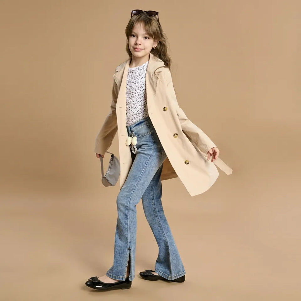 A young girl models a beige double-breasted trench coat over a white patterned top and blue flared jeans, styled with black flats and a small gray handbag against a solid beige studio background.