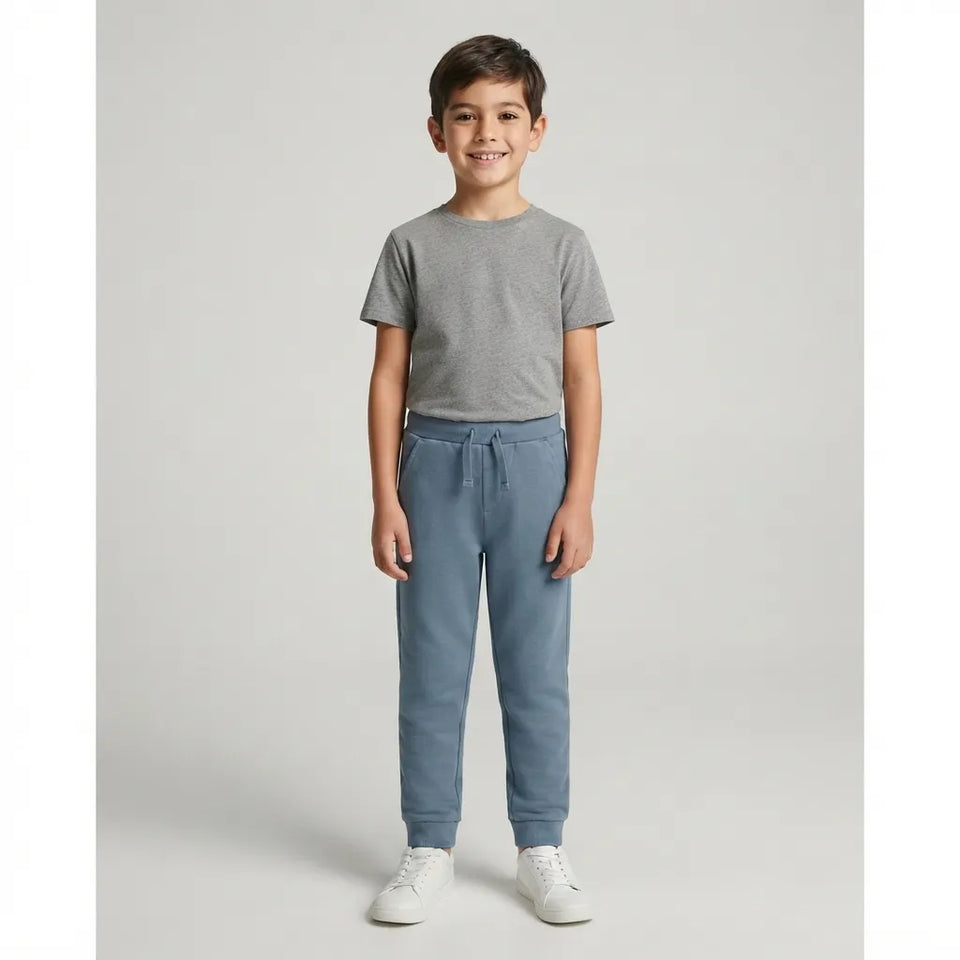 A young boy stands against a light gray studio background wearing a heather gray short-sleeve t-shirt, dusty blue sweatpants, and white sneakers.