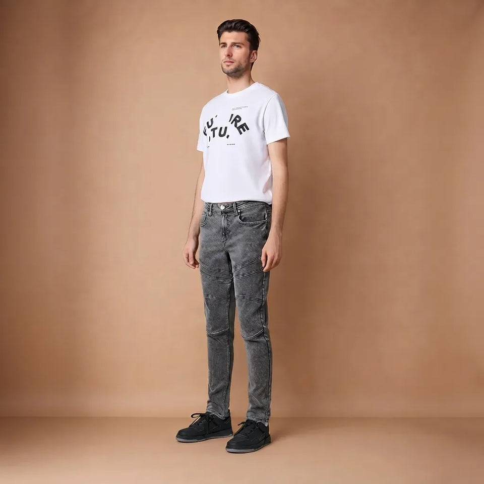 A man modeling a white graphic t-shirt, gray acid-wash skinny jeans, and black sneakers against a solid tan studio backdrop.