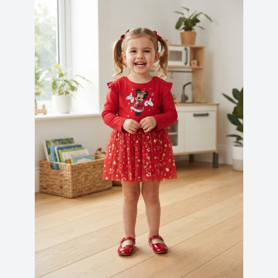 A young girl with blonde pigtails stands in a bright room wearing a red long-sleeve dress with a floral pattern and a cartoon character graphic on the chest, paired with red shoes.