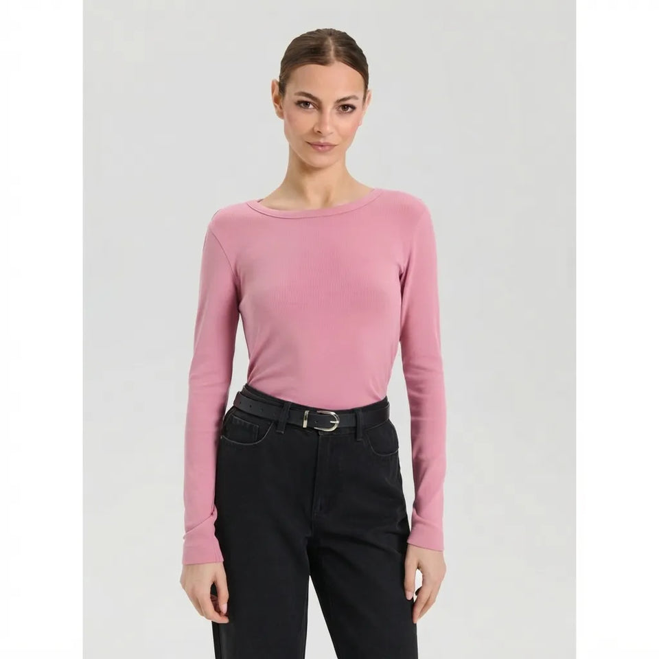 A woman modeling a long-sleeve crewneck top in dusty pink, paired with black jeans and a black belt, against a light gray studio background.