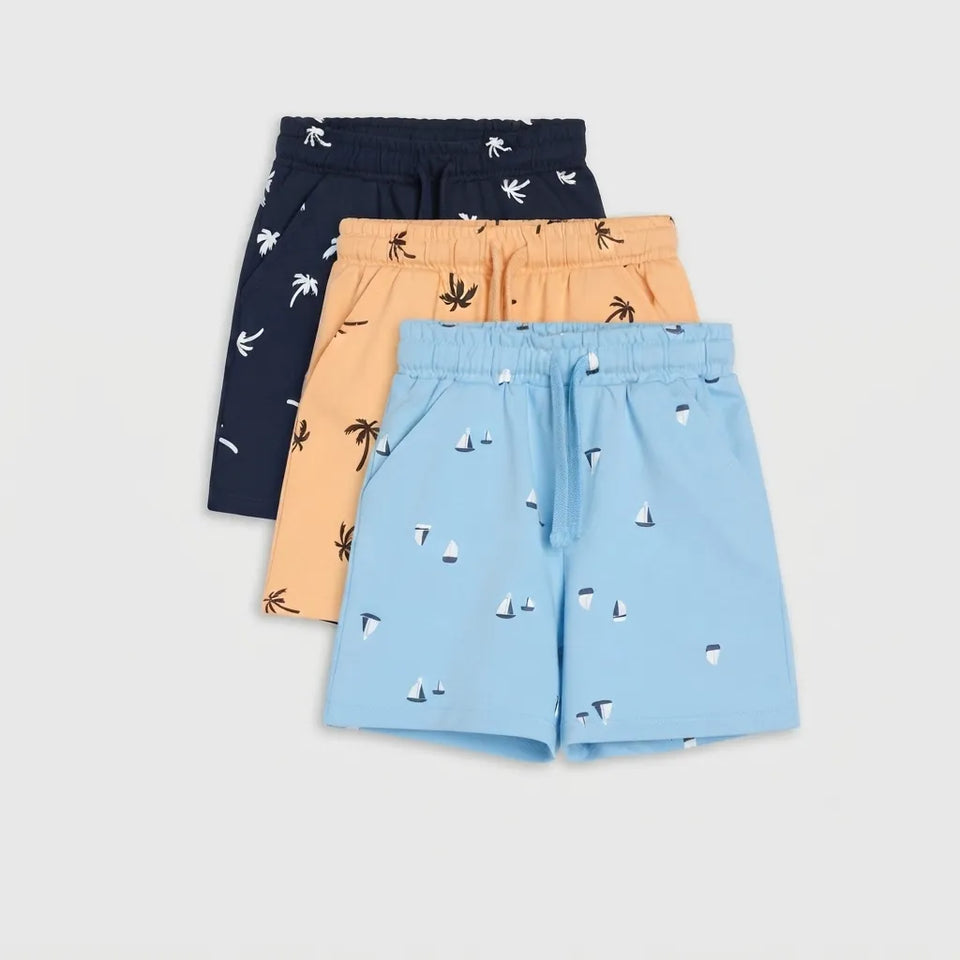 Three pairs of children's shorts in navy, peach, and light blue, each featuring a small tropical and nautical print, arranged in a fan shape on a light gray background.