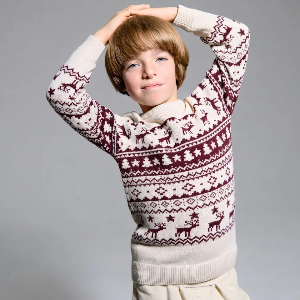 A young boy with light brown hair wearing a cream and burgundy Fair Isle knit sweater with reindeer patterns, posing with his arms raised against a solid gray studio background.