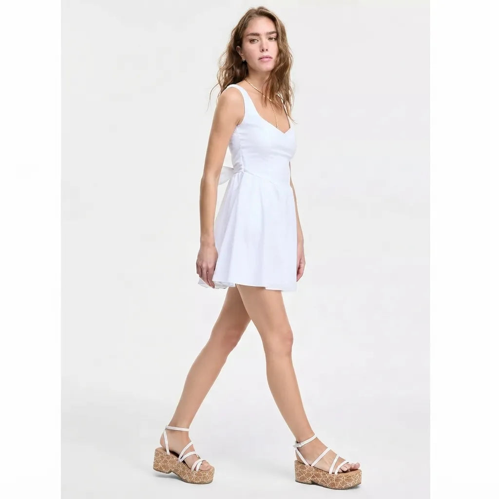 A woman modeling a white sleeveless mini dress with a V-neckline and a matching fabric tie at the waist, paired with white strappy platform sandals, against a plain white studio background.