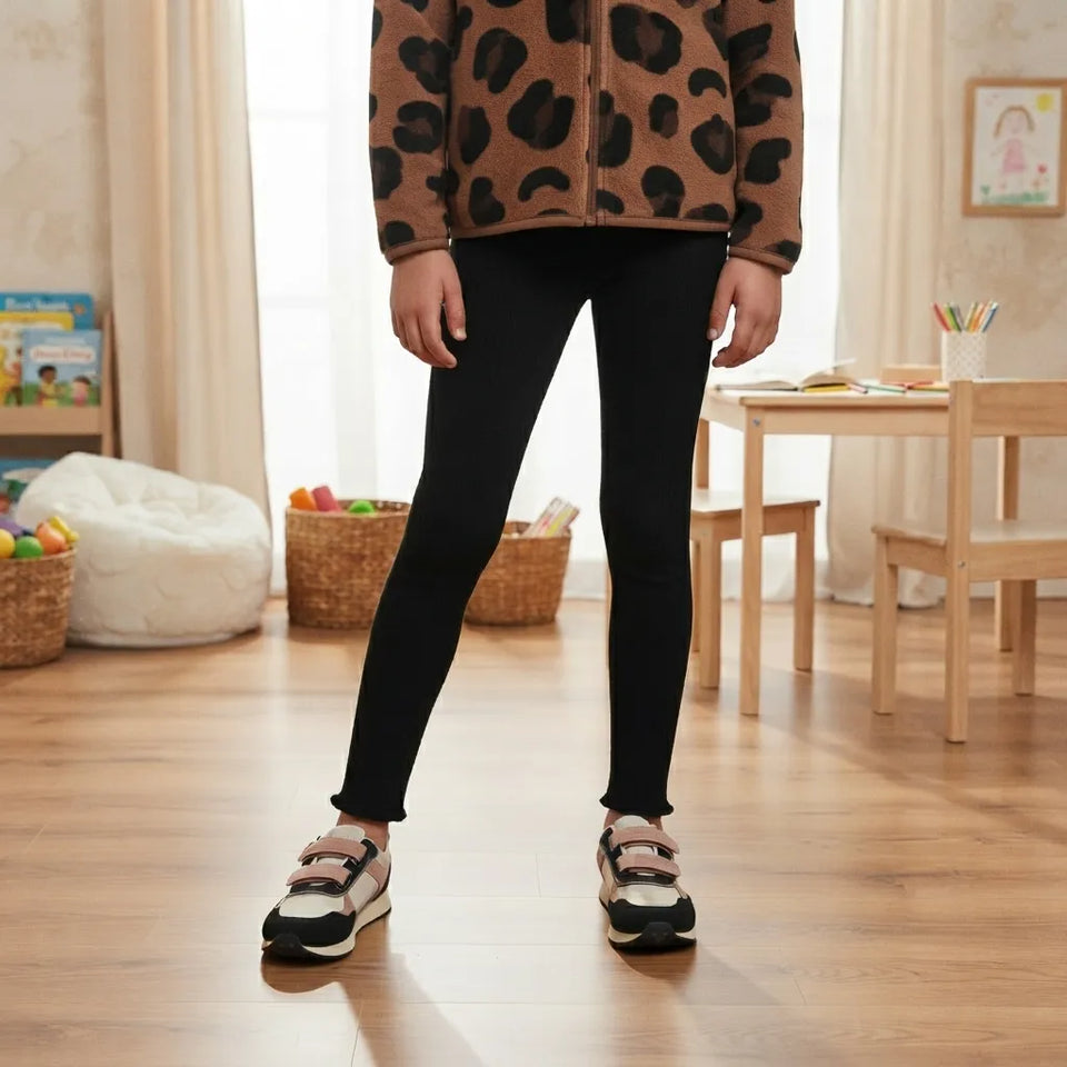 A child standing in a brightly lit room wearing black leggings, a brown fuzzy fleece jacket with black leopard spots, and black-and-white sneakers.