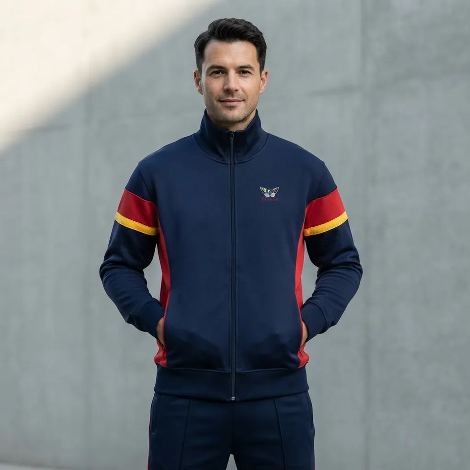A man wearing a navy blue zip-up track jacket with red and yellow sleeve stripes, standing against a gray concrete wall.