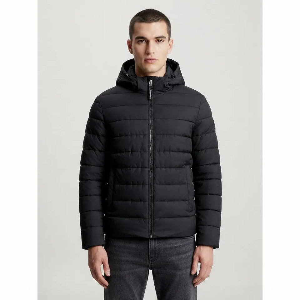 A man wearing a black hooded puffer jacket with horizontal quilting, shown from the waist up against a light gray studio background.