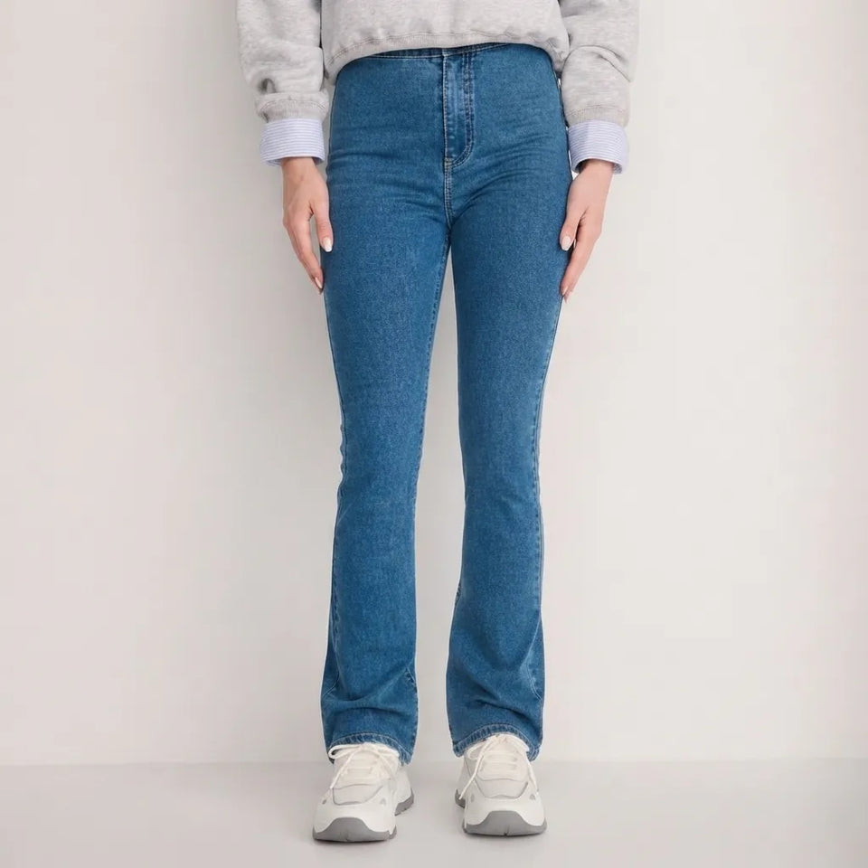 A person wearing medium-wash blue denim jeans with a straight-to-flare leg, paired with a light gray sweatshirt and white sneakers, standing against a plain light gray studio background.