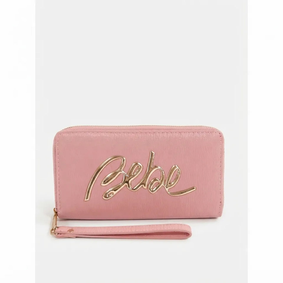 A pink zip-around wallet with a wristlet strap, featuring a metallic gold cursive 'Bebe' logo on the front, set against a light gray background.