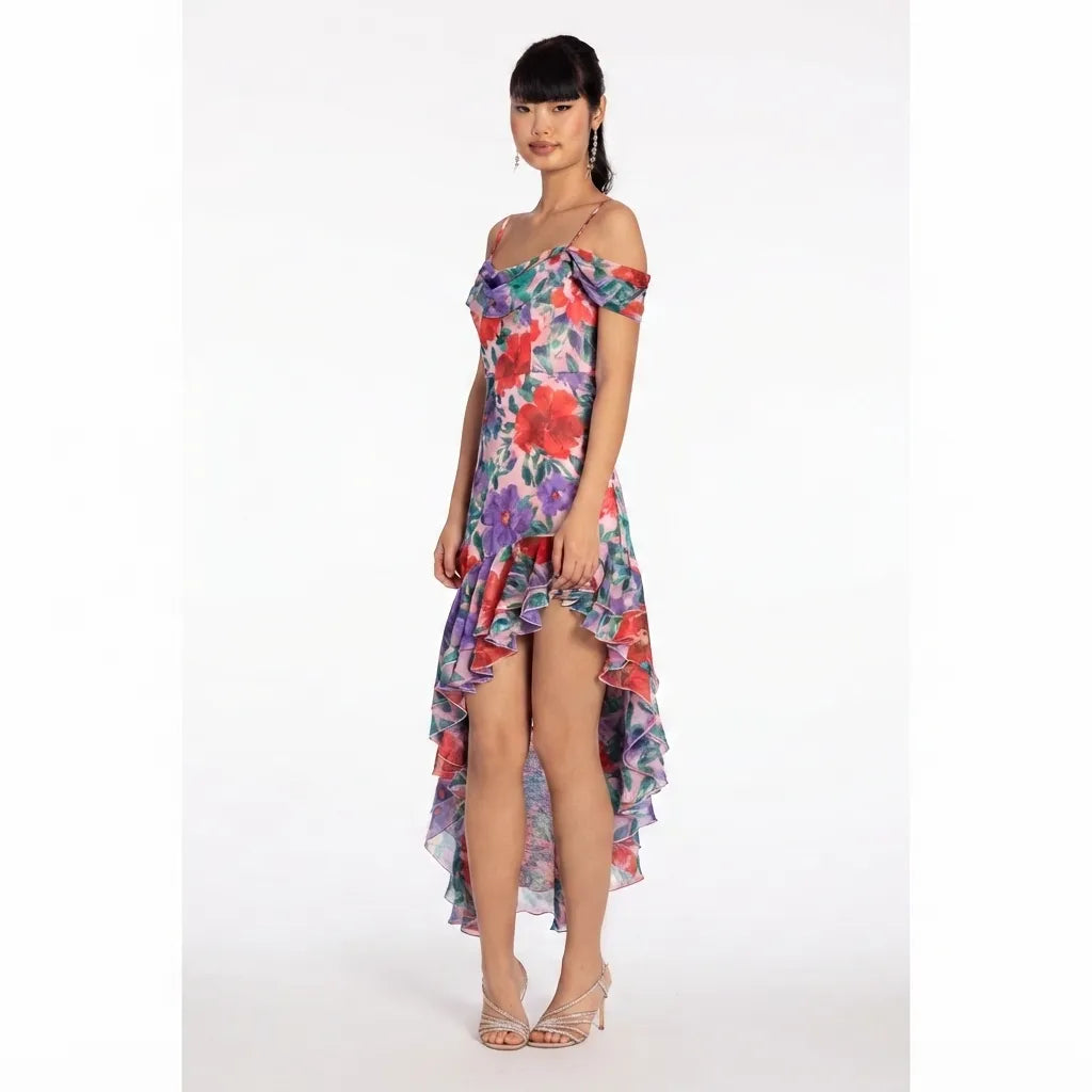 A woman modeling a floral off-the-shoulder high-low dress with a ruffled hem, photographed against a plain white studio background.