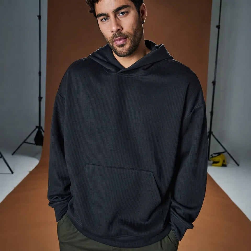 A man wearing a dark charcoal gray ribbed pullover hoodie with a front kangaroo pocket, standing in a studio setting with a brown backdrop.