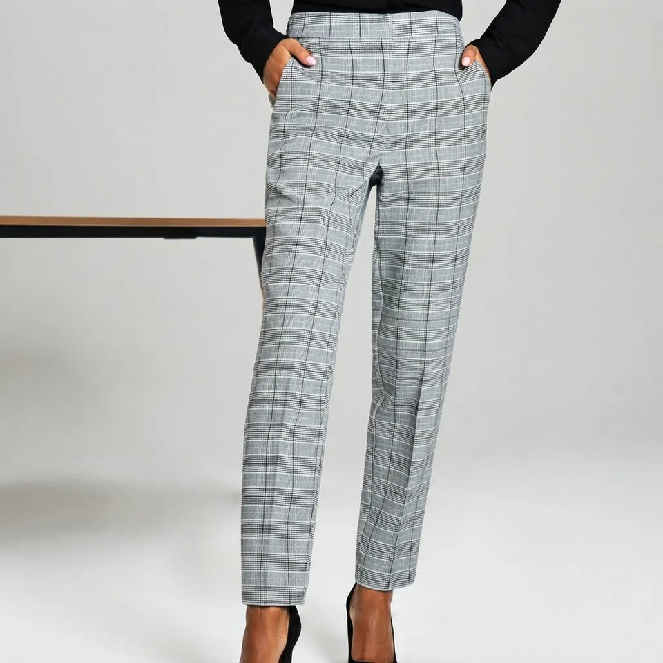 A person wearing slim, cropped plaid trousers in a light gray and white windowpane pattern, paired with a black long-sleeve top and black high heels, standing against a light gray studio background.