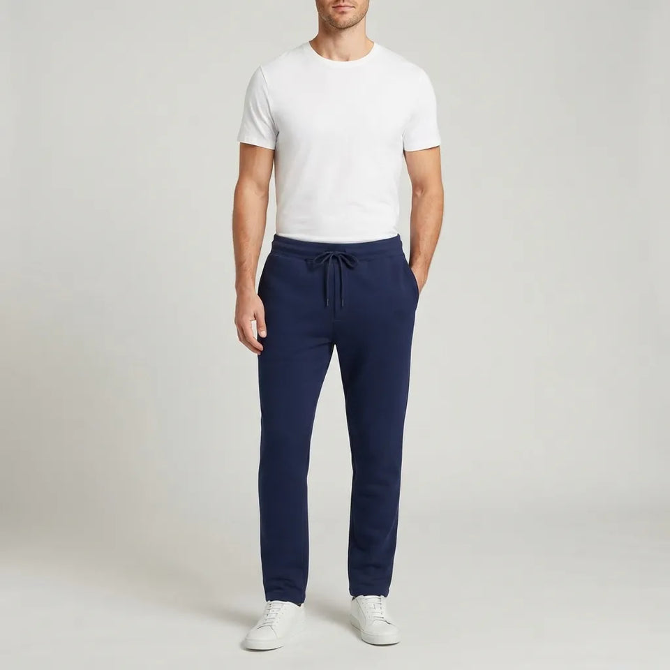 A man modeling navy blue drawstring sweatpants with a white t-shirt and white sneakers, standing against a light gray studio background.
