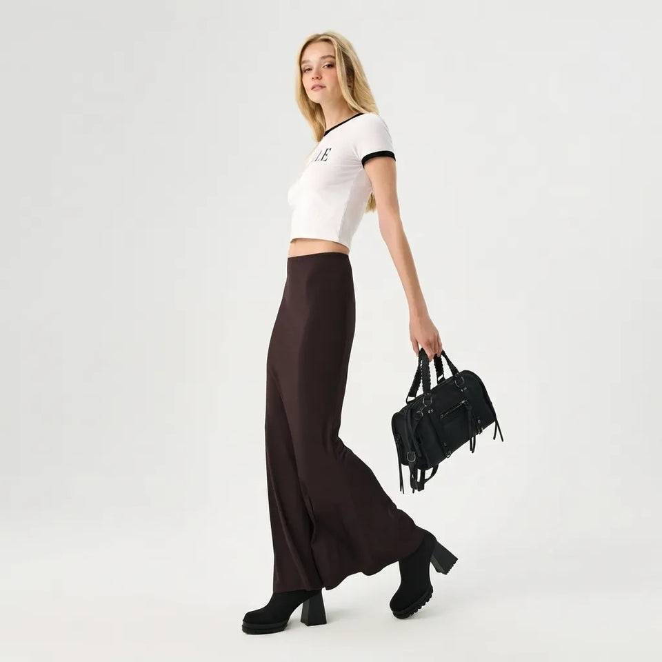 A woman modeling a white short-sleeve crop top with black trim, dark brown wide-leg pants, black ankle boots, and carrying a black handbag against a light gray studio background.