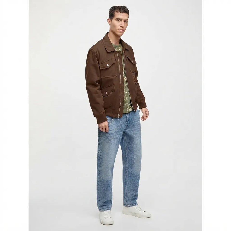 A man modeling a brown utility jacket over a green patterned shirt, paired with blue jeans and white sneakers, against a plain light gray studio background.
