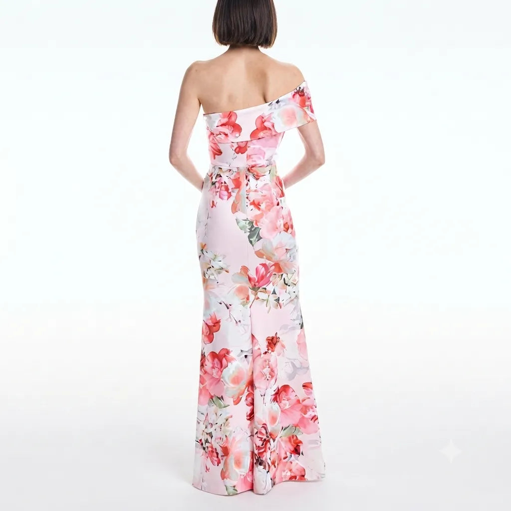 A woman shown from the back wearing a one-shoulder off-the-shoulder maxi dress with a pink and red floral print on a white base, set against a plain white studio background.