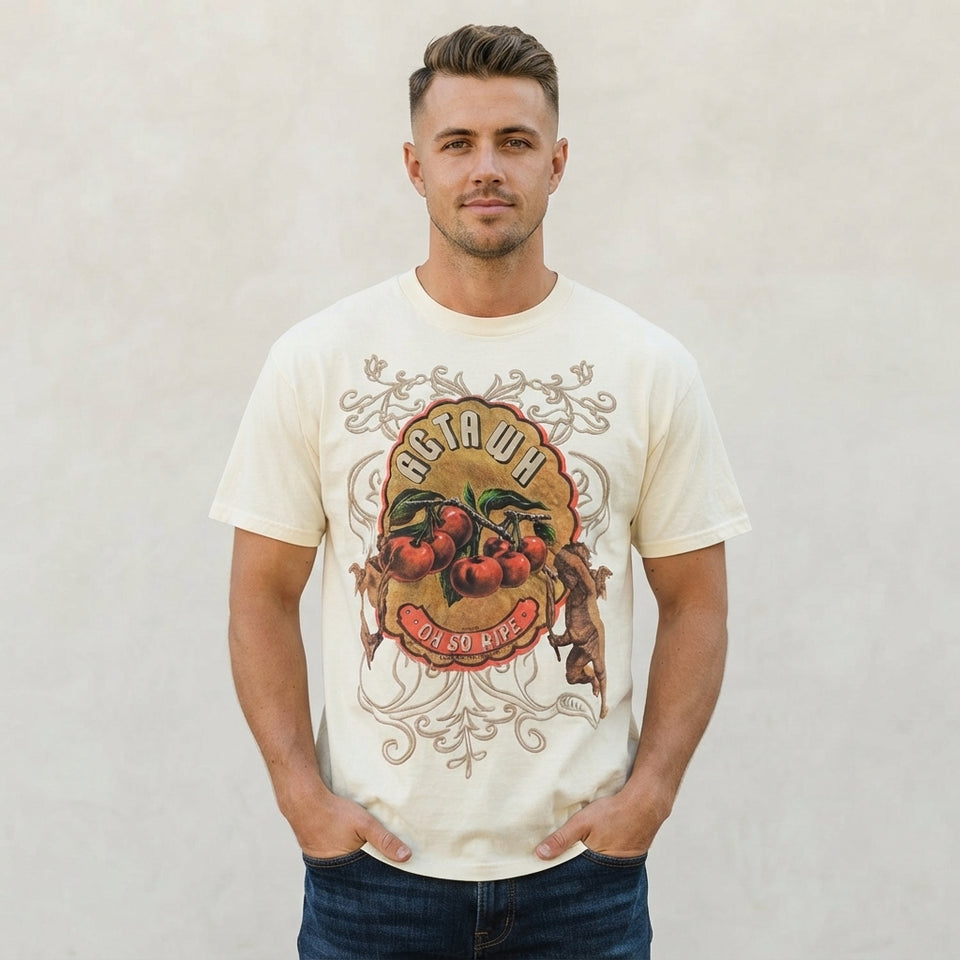 A man wearing a cream-colored graphic t-shirt with a vintage-style fruit and floral design on the front, paired with dark blue jeans, standing against a neutral textured wall.