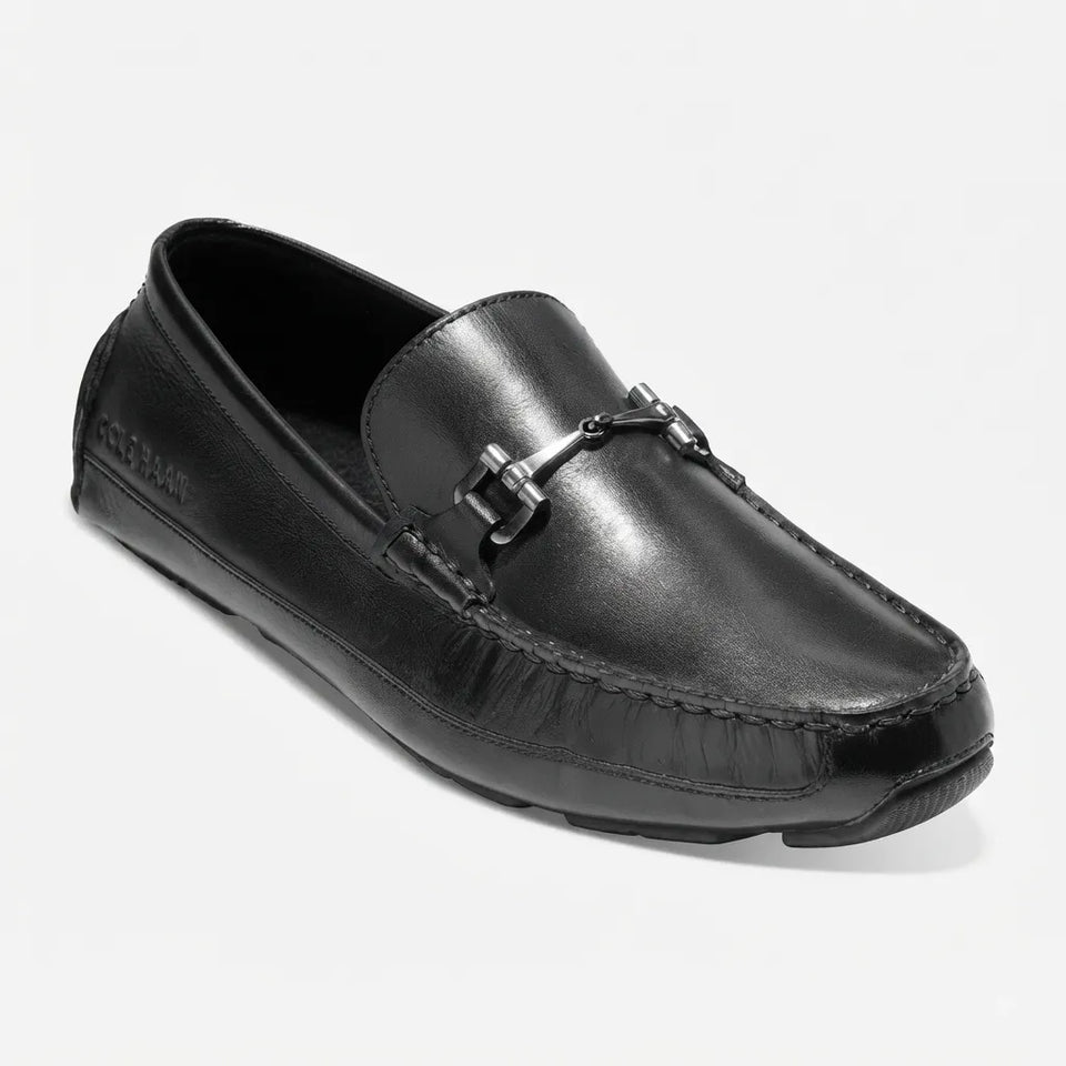 A black leather driving loafer with a silver-tone horsebit detail on the vamp, photographed from a three-quarters angle against a light gray background.