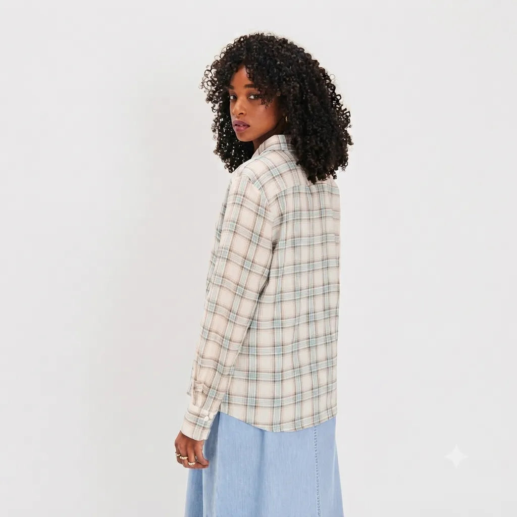 A woman with curly black hair is shown from a side-back angle wearing a cream-colored long-sleeve plaid button-up shirt with thin green and blue lines, paired with a light blue skirt, against a plain white studio background.