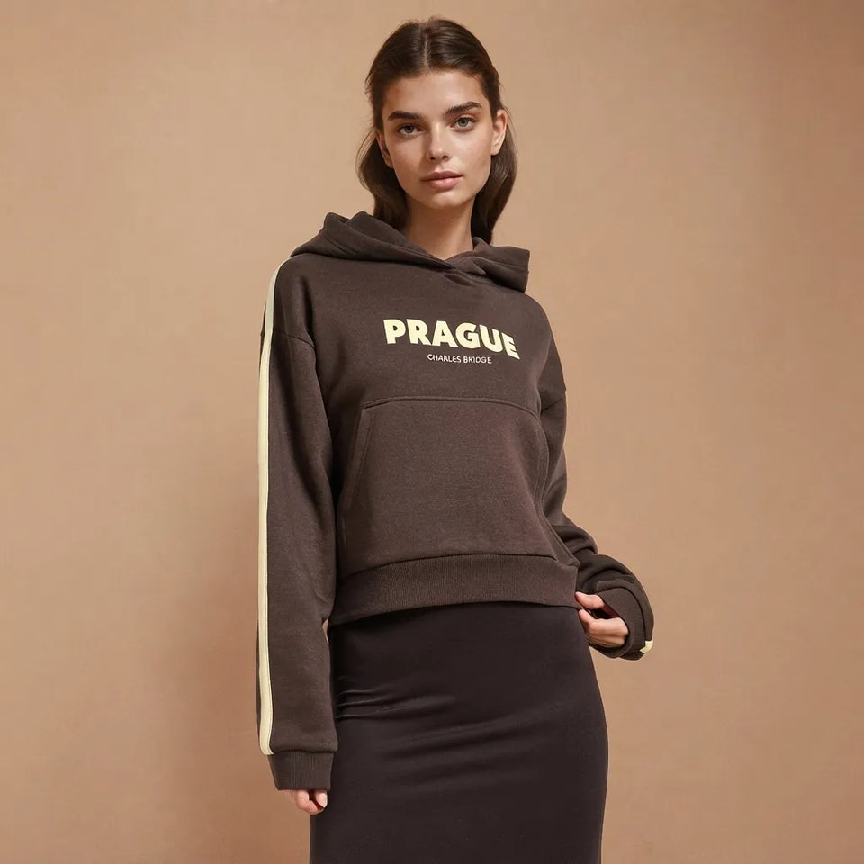 A woman modeling a dark brown cropped hoodie with 'PRAGUE' printed in yellow across the chest and a cream stripe down the sleeve, paired with a black skirt, against a solid beige background.