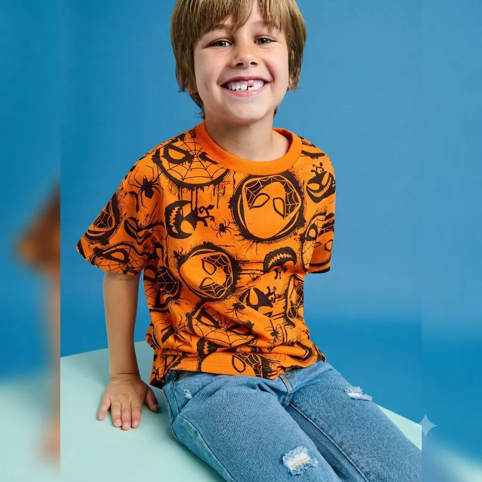 A young boy with light brown hair smiling, wearing a bright orange short-sleeve t-shirt with a black spider-themed graphic print and light blue jeans, sitting against a solid blue background.
