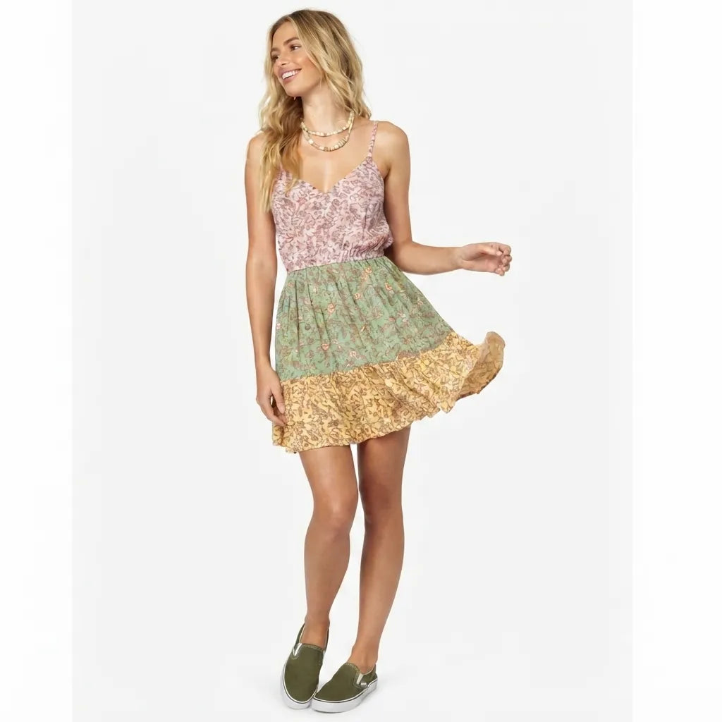 A woman modeling a sleeveless mini dress with a pink floral bodice, a sage green floral waist, and a gold glittery ruffled hem, paired with olive green slip-on shoes against a plain white background.