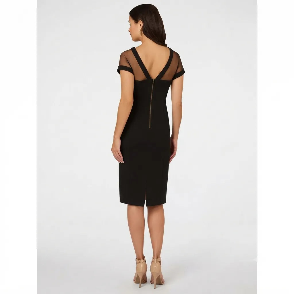 A woman modeling a black knee-length dress from the back, featuring sheer mesh shoulder straps, a V-neckline, and a central zipper, paired with beige high-heeled sandals against a light gray studio background.