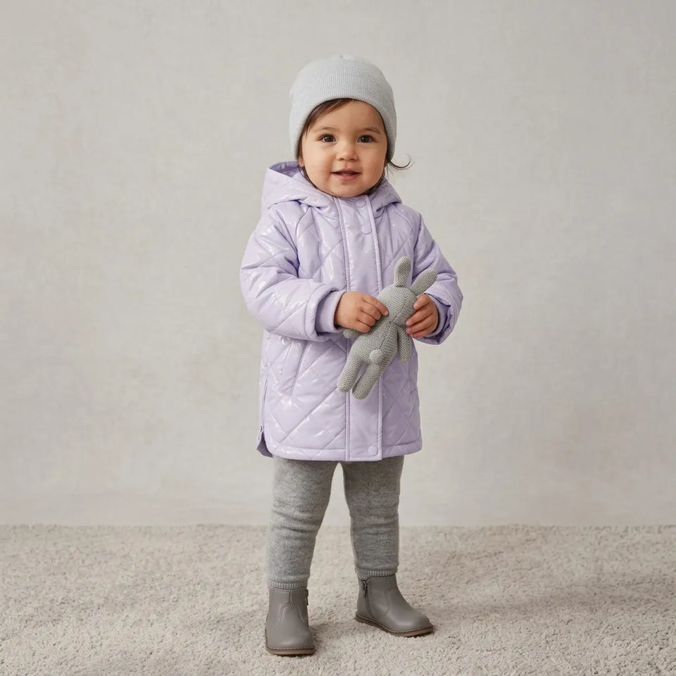 A toddler stands on a light carpet wearing a light lavender quilted hooded jacket, gray leggings, gray boots, and a light gray beanie, while holding a gray stuffed bunny.