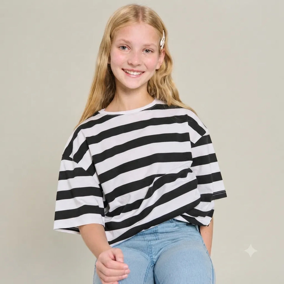A young girl with blonde hair wearing a black and white horizontally striped short-sleeve t-shirt and light blue jeans, posing against a solid light gray background.