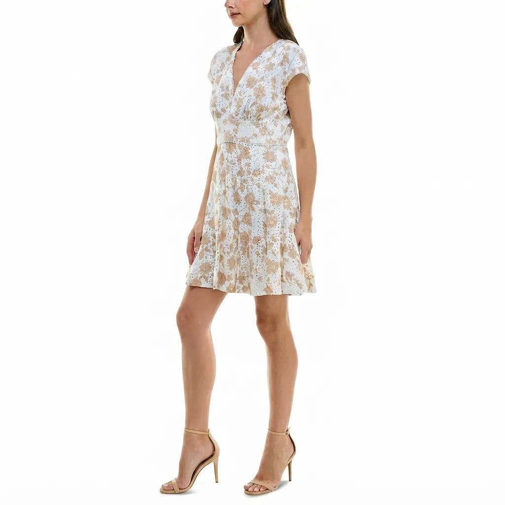 A woman modeling a short white wrap-style dress with a beige floral pattern, cap sleeves, and a V-neckline, paired with beige high-heeled sandals against a plain white background.