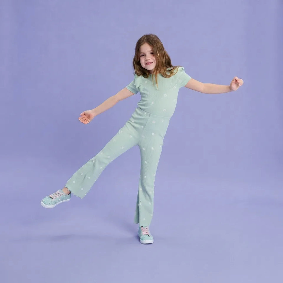 A young girl with light brown hair is posing in a mint green short-sleeve jumpsuit with small white polka dots, paired with matching mint sneakers, against a solid lavender studio backdrop.