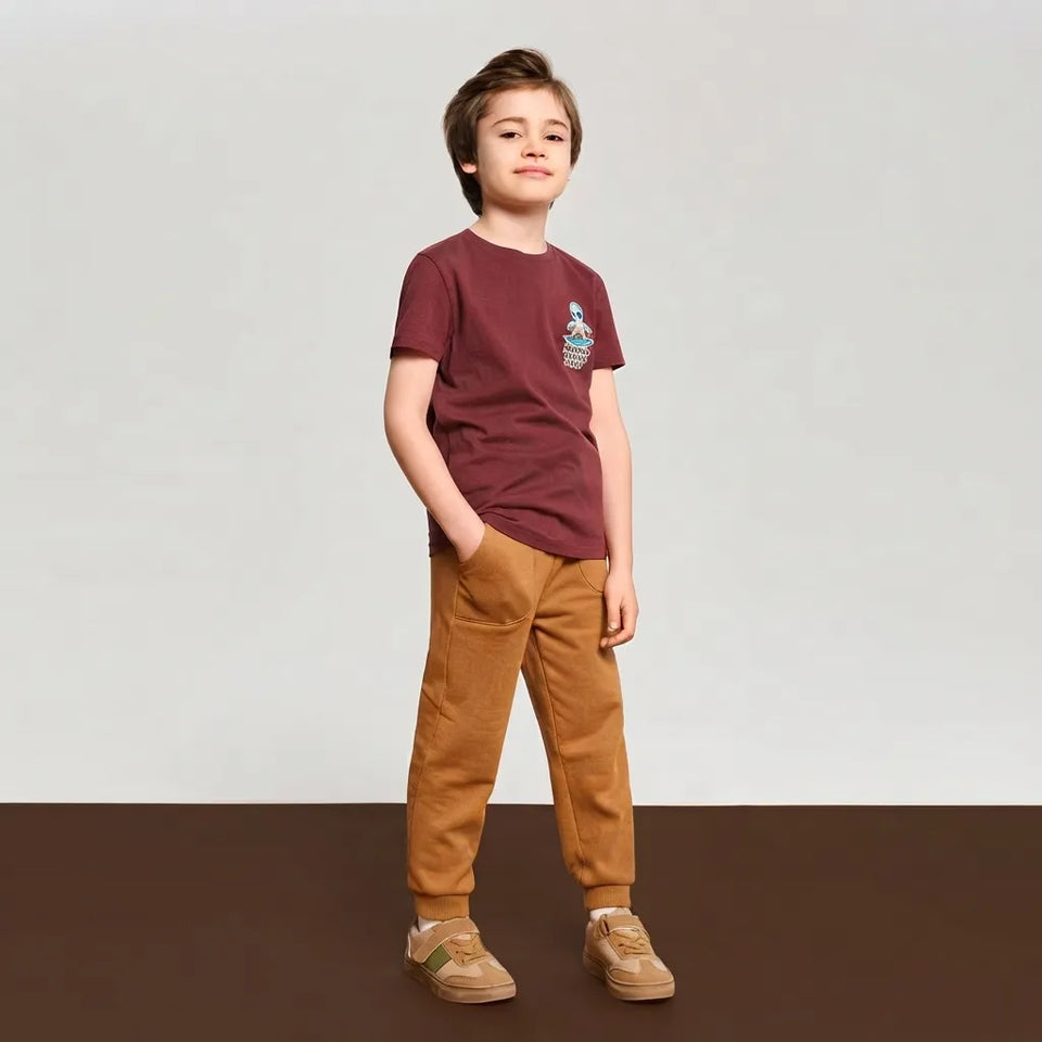 A young boy stands against a neutral studio backdrop wearing a short-sleeve crewneck t-shirt in maroon with a small graphic on the left chest, paired with mustard brown corduroy pants and beige sneakers.