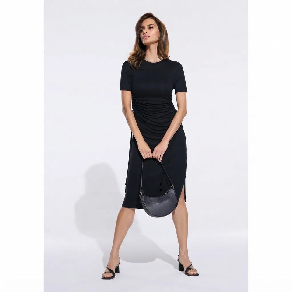 A woman modeling a black short-sleeve midi dress with ruching at the waist, holding a dark gray crescent-shaped handbag against a light gray studio background.