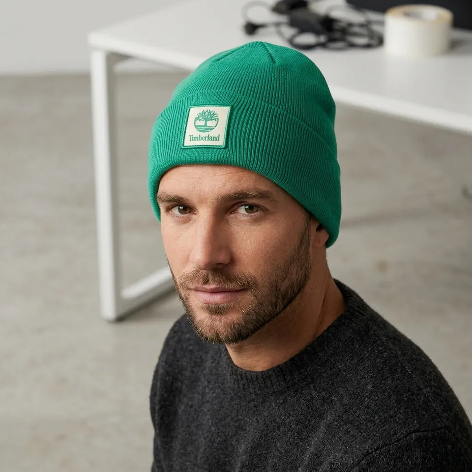 A man wearing a bright green Timberland knit beanie with a white brand patch on the front, photographed in a modern indoor setting with a white table in the background.