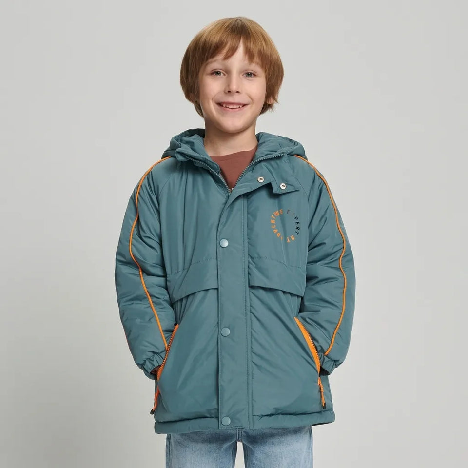 A smiling young boy with light brown hair stands against a light gray studio background wearing a teal hooded puffer jacket with orange piping and zipper accents, a brown shirt underneath, and blue jeans.