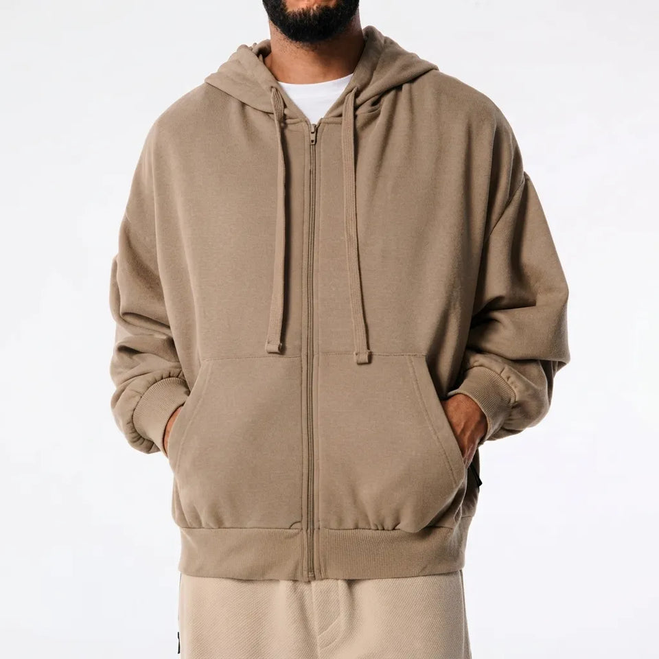 A man wearing a taupe-brown zip-up hoodie with drawstrings and a front kangaroo pocket, layered over a white t-shirt and beige sweatpants, against a plain white background.