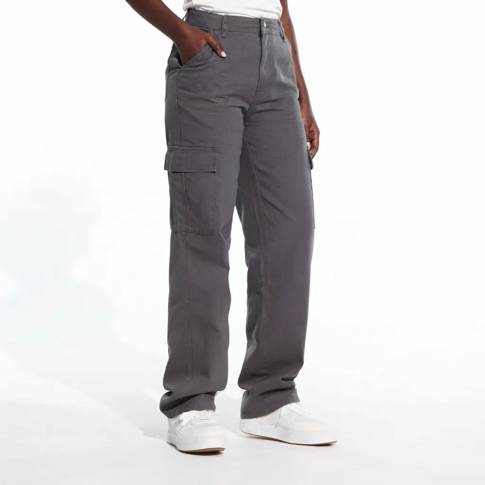 A person wearing dark gray cargo pants with side flap pockets, paired with a white top and white sneakers, shown from the waist down against a white studio background.