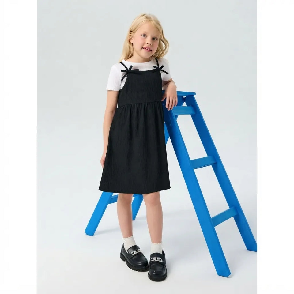 A young girl stands on a small blue step stool in a studio setting, wearing a black pinafore-style dress over a white short-sleeve top, with black loafers and white socks.