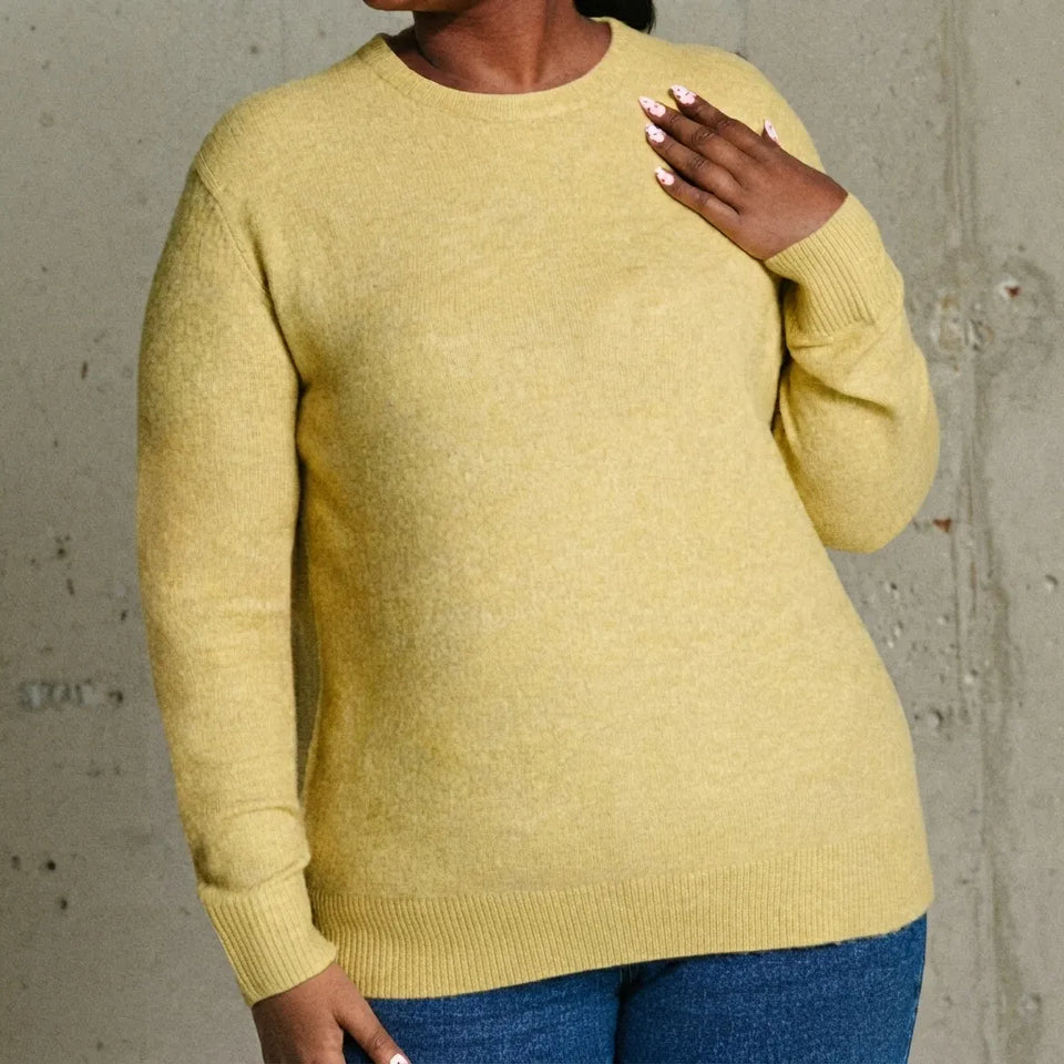 A person wearing a soft heathered pale yellow knit sweater with blue jeans, standing against a textured concrete wall.