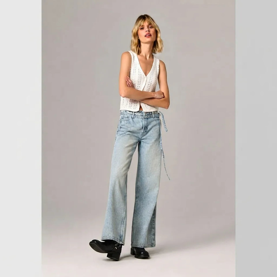 A model stands against a light gray studio background wearing a white sleeveless crochet vest, light-wash wide-leg jeans with a blue denim tie, and black shoes.