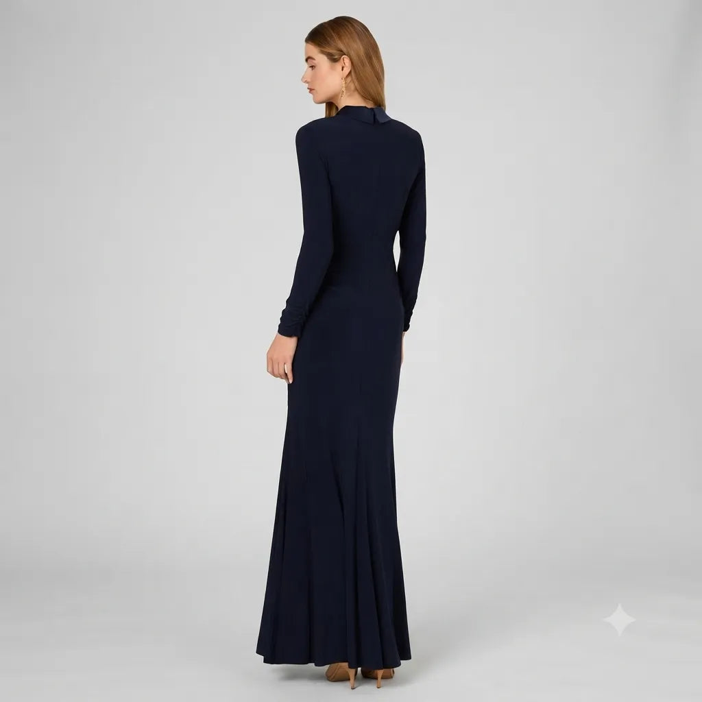 A woman modeling a long-sleeved, high-neck, floor-length navy blue dress, shown from a rear three-quarter view against a light gray studio background.