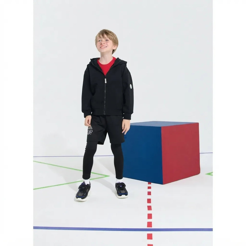 A young boy wearing a black zip-up hoodie, black shorts, and black leggings stands next to a blue and red cube on a white studio floor with colored lines.
