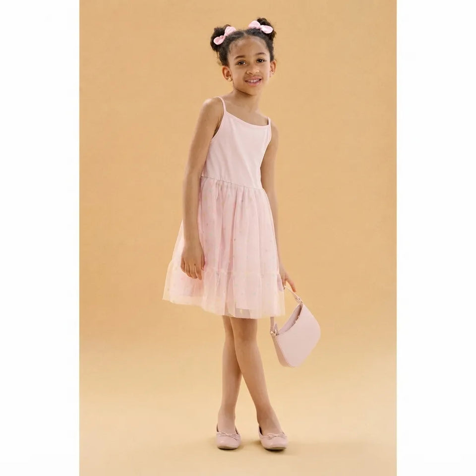 A young girl with her hair in two buns wearing a light pink sleeveless dress and holding a matching light pink handbag, standing against a solid beige background.