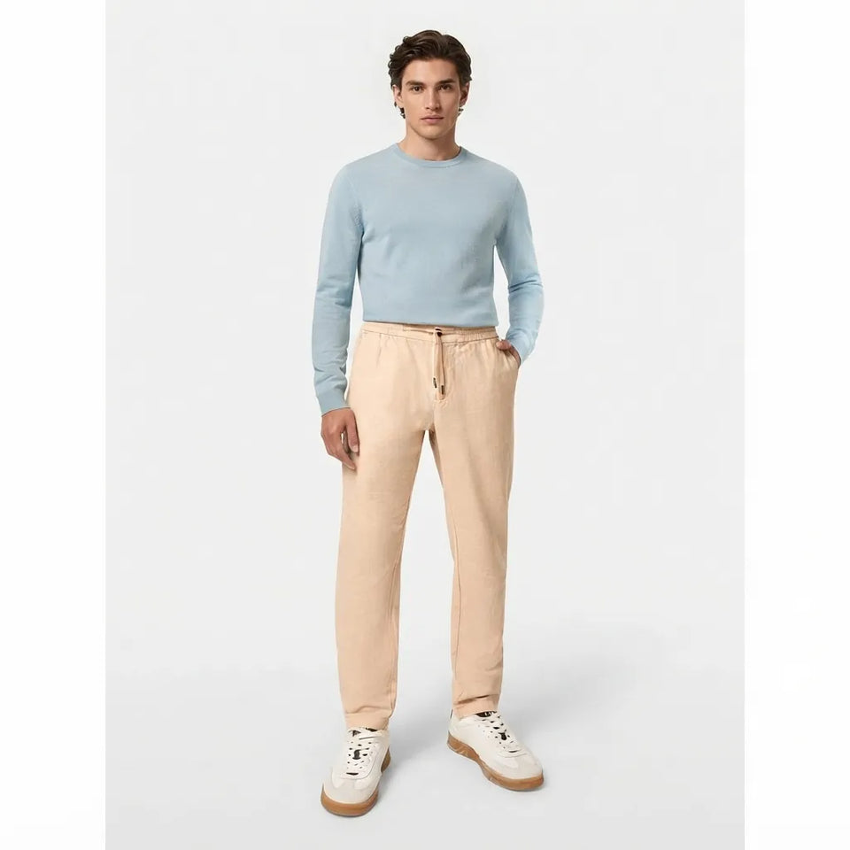 A man modeling a light blue crewneck sweater and beige drawstring sweatpants with white sneakers, standing against a light gray studio background.