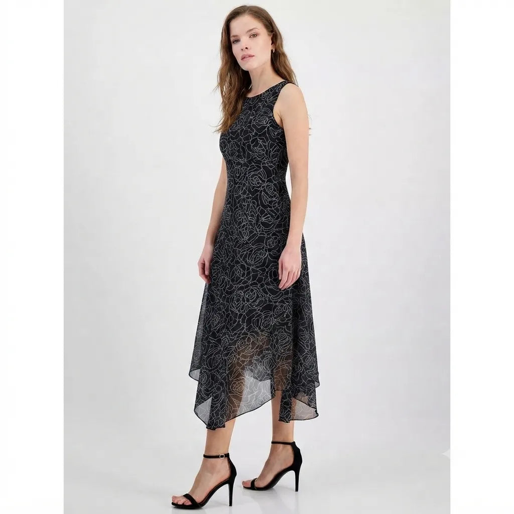 A woman modeling a sleeveless black midi dress with a subtle tonal floral lace pattern and a high-low hemline, paired with black strappy heels against a light gray studio background.