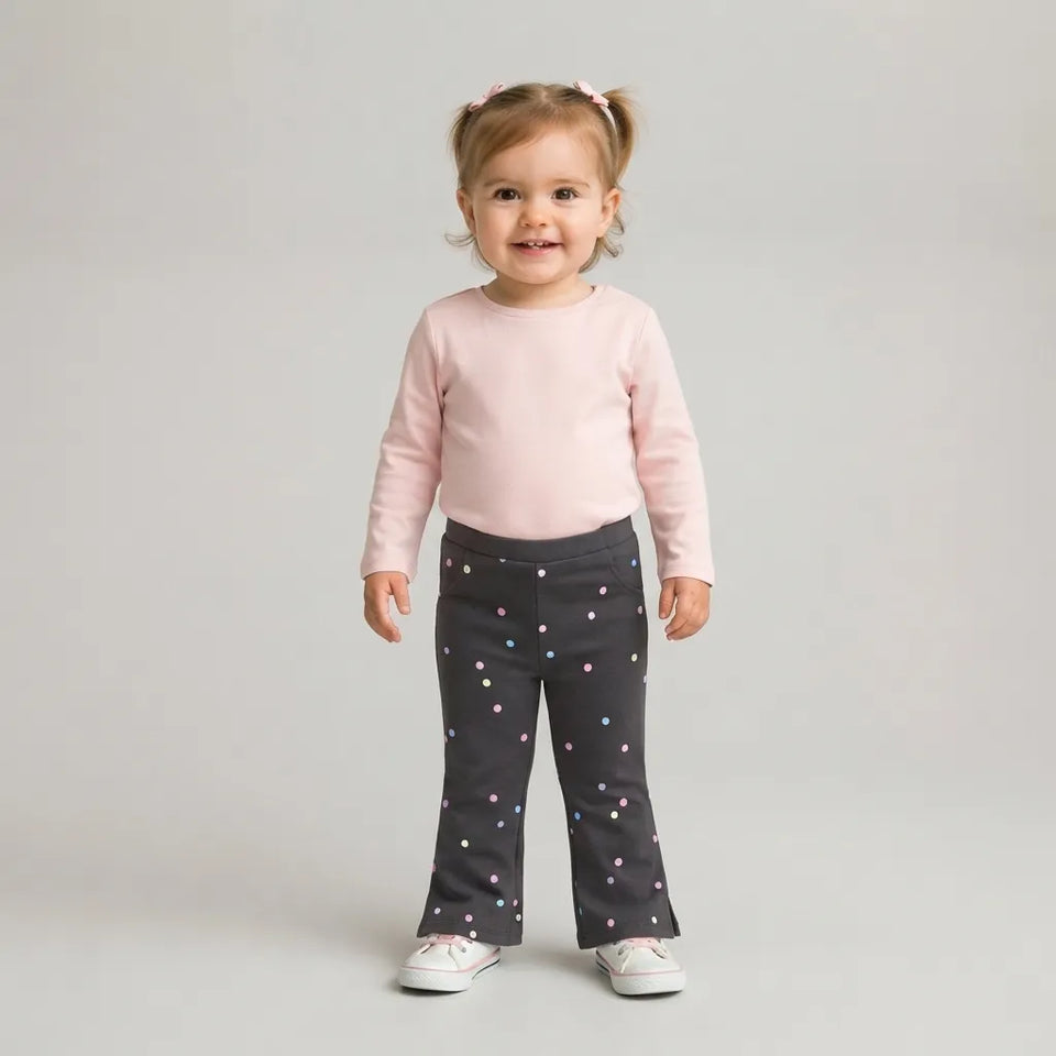 A toddler girl standing against a light gray studio background, wearing a light pink long-sleeve top and dark gray flared pants with small pink, blue, and white polka dots. She is smiling and looking at the camera.