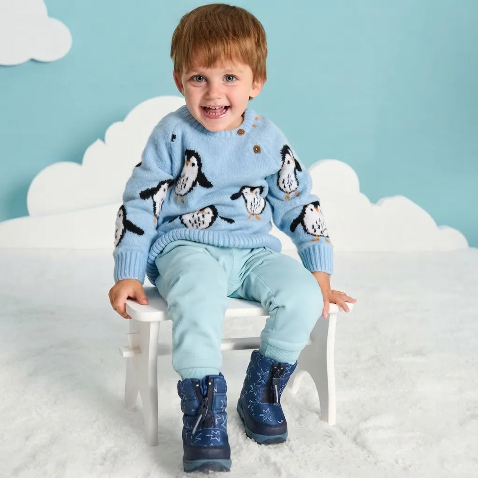 A smiling young boy sits on a small white stool wearing a light blue knit sweater with penguin patterns, matching light blue pants, and dark blue snow boots against a light blue background with white cloud cutouts.