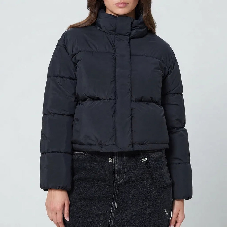 A model wearing a cropped black puffer jacket with a high stand-up collar and horizontal quilting, paired with a dark textured skirt, against a light gray studio background.