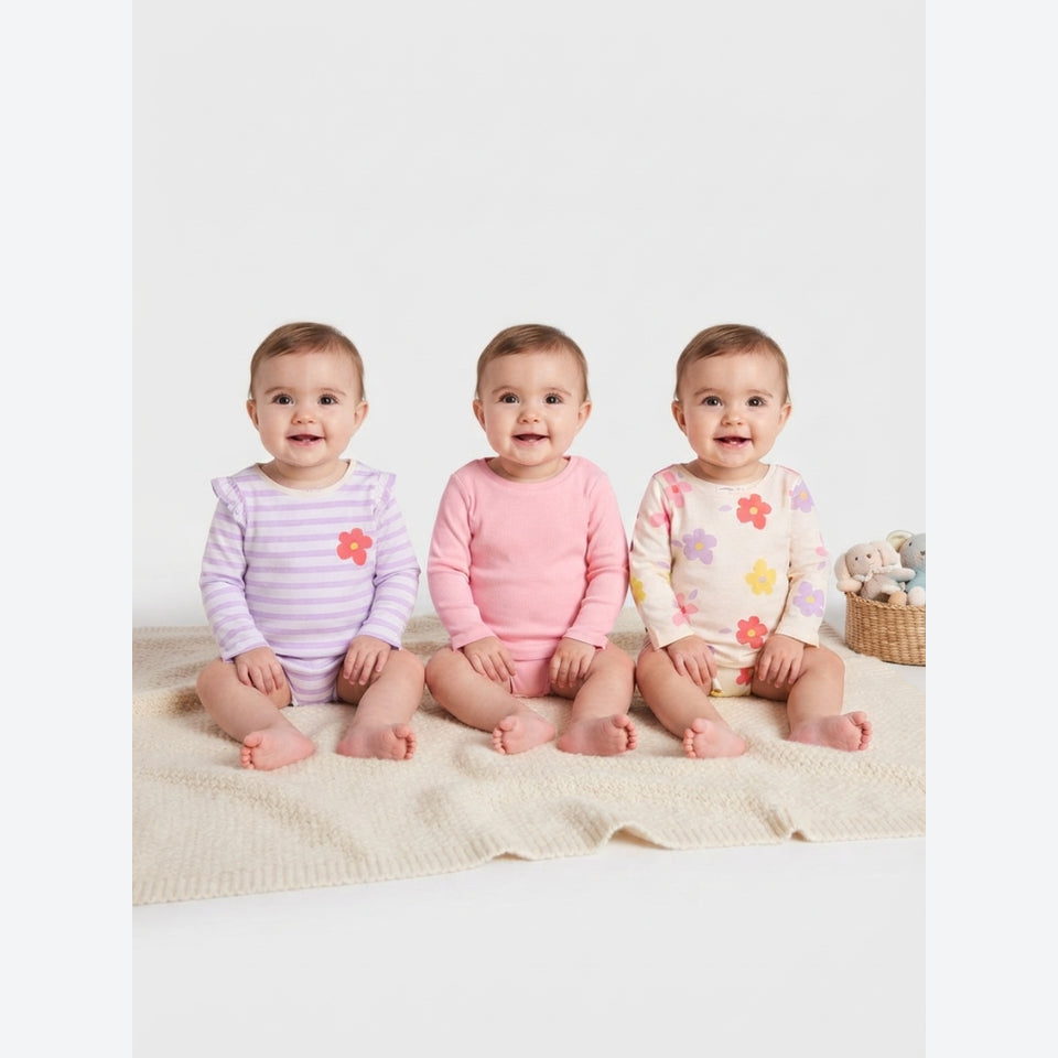 Three babies sitting side by side on a cream knit blanket, wearing long-sleeve baby bodysuits. The baby on the left wears a light purple and white striped bodysuit with a pink flower on the chest.